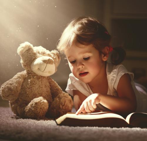Niña pequeña acostada en una alfombra, señalando con su dedo un libro abierto que lee con atención. A su izquierda, un oso de peluche sentado la acompaña. Un haz de luz cálida ilumina a ambos desde arriba, creando una atmósfera mágica y de aprendizaje.