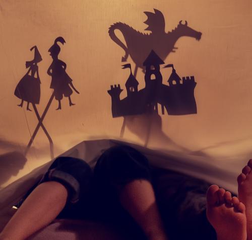Fotografía de las piernas de una persona acostada dentro de una carpa de sábanas con luz cálida. Sobre la tela se proyectan sombras chinescas de figuras infantiles: una niña, un niño con espada, un castillo y un dragón en la parte superior.