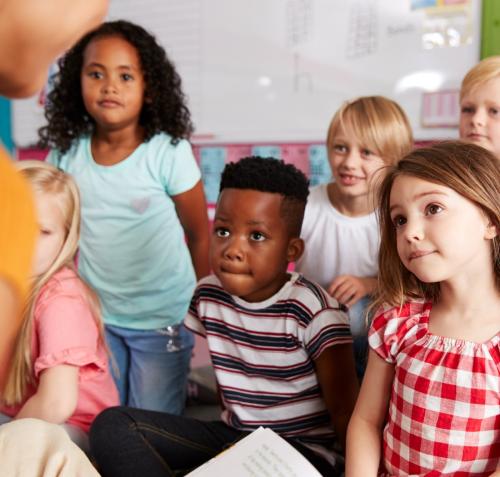 Fotografía de cinco niños de diversas etnias sentados en el suelo de un aula escolar. Miran con mucha atención hacia un adulto, parcialmente visible, que sostiene un libro. Al fondo hay un pizarrón. Las expresiones de los niños reflejan gran interés y concentración.