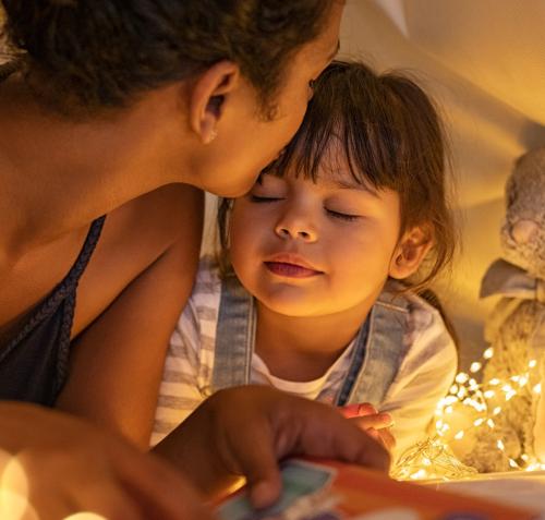 Una mujer besa tiernamente la frente de una niña que sonríe con los ojos cerrados. Comparten la lectura de un libro en un espacio acogedor, bajo una sábana iluminada por cálidas luces de hadas.