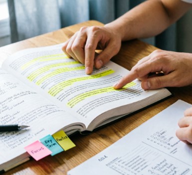 Manos de un estudiante sobre un libro abierto con líneas resaltadas en amarillo; un dedo señala el texto. Notas adhesivas rosa, azul y amarilla dicen 'Focus', 'Key Term', 'Practice'. Junto al libro, un bolígrafo y una hoja de ejercicios sobre un escritorio de madera con luz natural.
