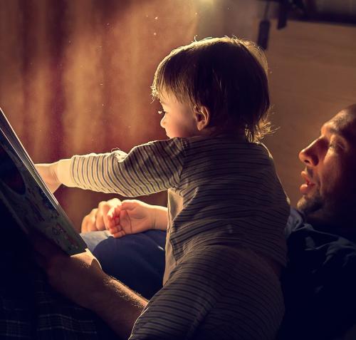 Un hombre y un niño pequeño comparten la lectura de un libro en un ambiente oscuro iluminado por una luz cálida. El niño, con pijama a rayas, señala la página abierta mientras el adulto sostiene el libro y lee, creando un momento íntimo.