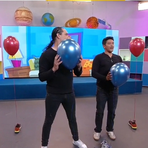 Dos hombres en un colorido set de TV con planetas colgantes. El de la izquierda oculta su rostro con un gran globo azul; el derecho sonríe con otro globo azul en el pecho. A los lados, globos rojos atados al suelo. De fondo hay una pantalla con dibujos infantiles.