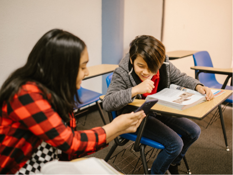Una niña de pelo largo y un niño, sentados en sus pupitres, miran riendo algo en el celular de ella