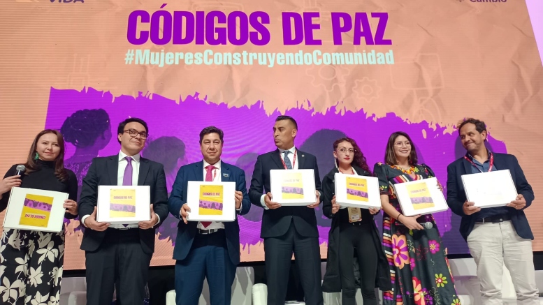 Foto de cuatro hombres y tres mujeres sosteniendo una caja, con un letrero al fondo que dice Códigos de paz en letra de color lila