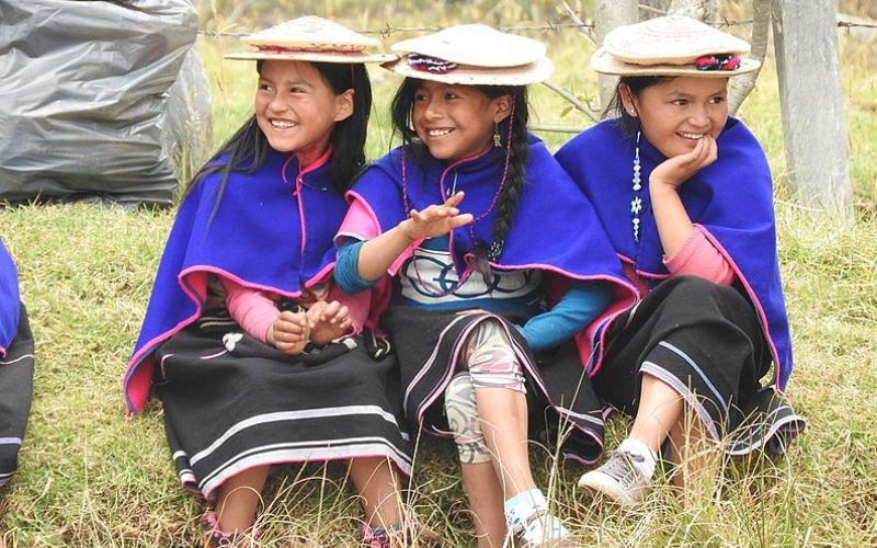 Tres niñas indigenas colombianas con trajes típicos, falda negra, ruana azul, sombrero blanco, sentadas en el pasto sonrien