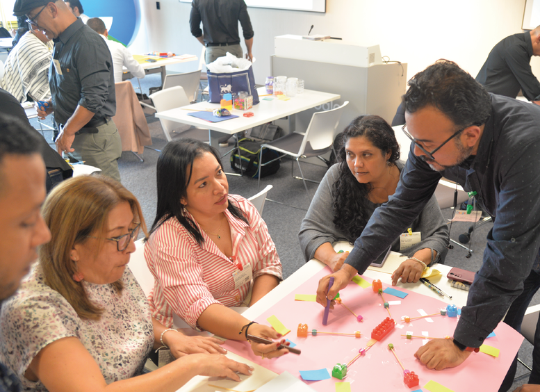 Varios docentes, hombres y mujeres sentados trabajando alrededor de una mesa y un hombre les da indicaciones