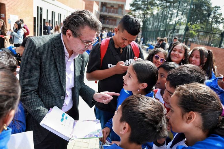 Foto del ministro Alejandro Gaviria hablando con niños en el patio de un colegio, mientras sostiene un cuaderno