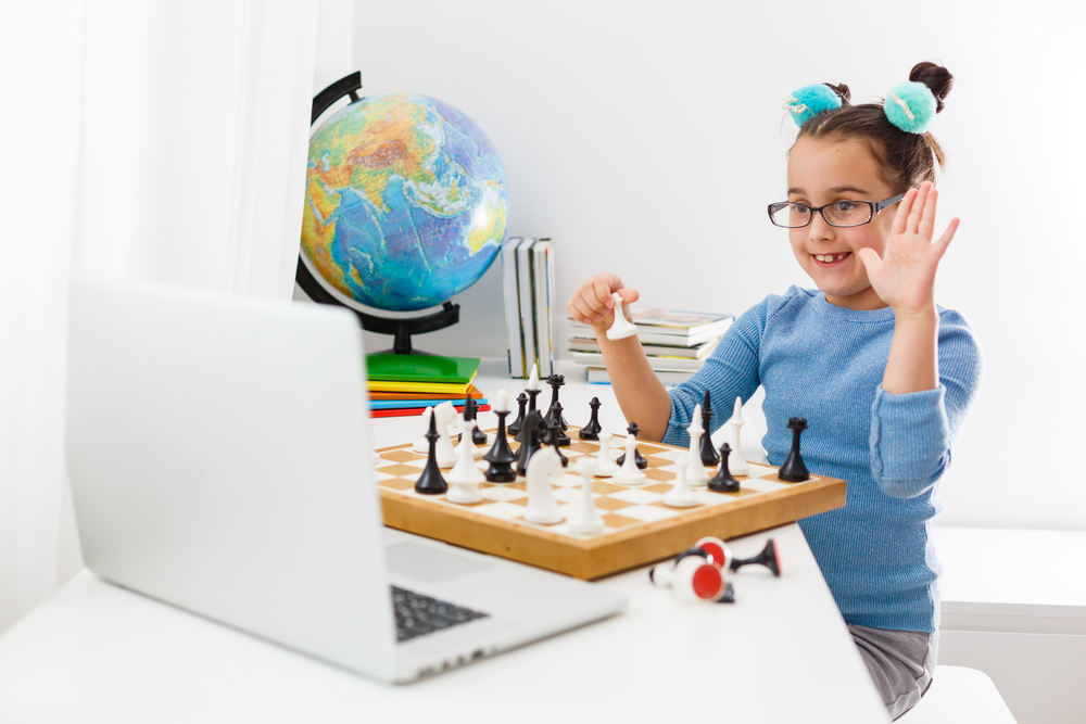 Fotografía de una niña jugando ajedrez frente a un computador, al fondo un mapamundi