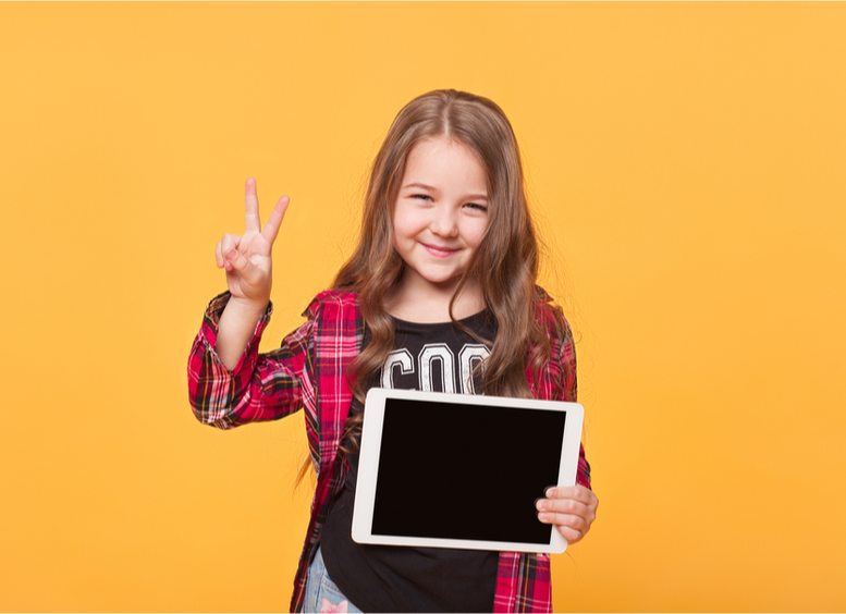 Niña de pelo largo sonriente, camisa a cuadros, sostiene una tableta en la mano y hace el signo de victoria con la mano