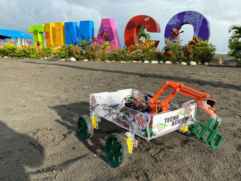 Foto de prototipo robótico sobre el piso de tierra, y al fondo la palabra TUMACO en letras de colores