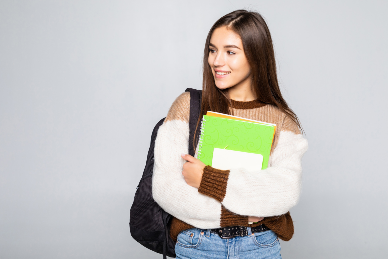 Foto de mujer joven estudiante universitaria de pelo negro sonriendo, que abraza cuadernos y lleva un morral