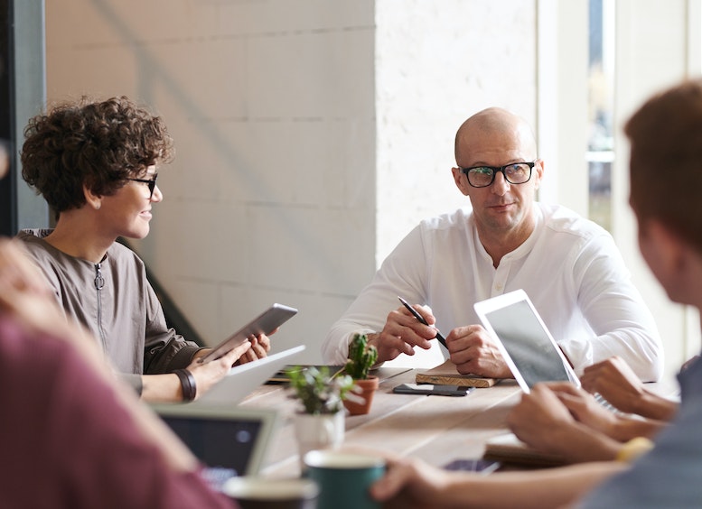 Cuatro personas con textos, tabletas, y libros conversan en una mesa, observando al que es el profesor