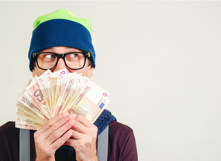 Joven con gorro y gafas sosteniendo un manojo de billetes que le tapan media cara, y una mirada de sorpresa