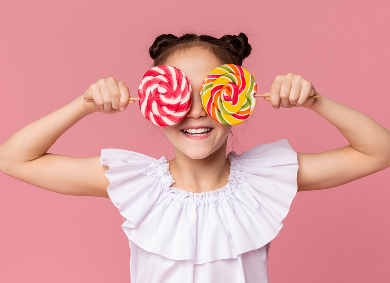 Niña vestida de blanco cubriendo sus ojos con dos colombinas grandes de colores, fondo rosa