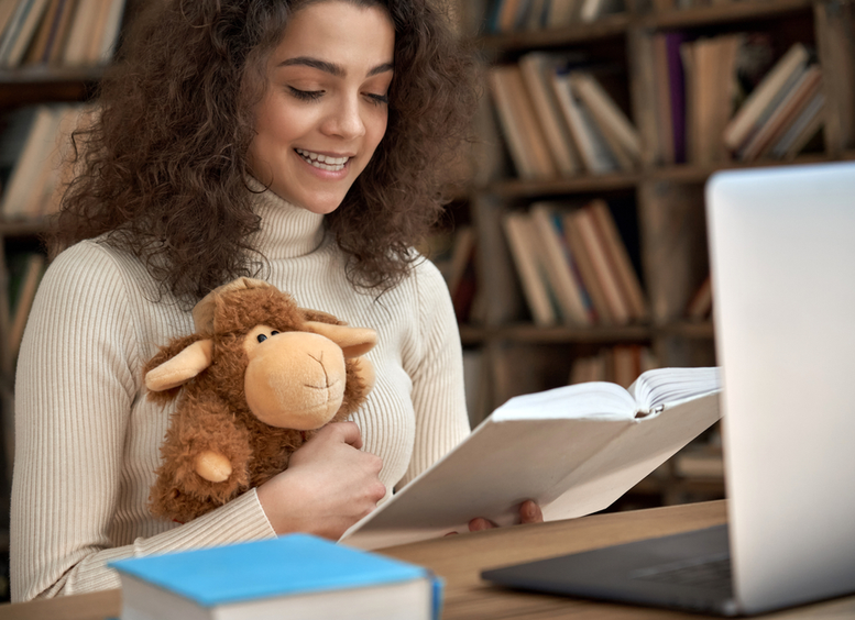 Una profesora joven, trigueña de pelo negro, sentada con una biblioteca de fondo, sostiene a una oveja cafe de juguete, y lee un libro de cuentos frente a una computadora portatil