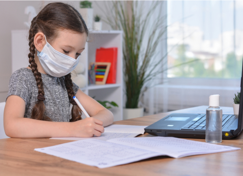 Una colegiala con una máscara médica está haciendo sus deberes en una laptop en casa o estudiando en clase en una lección en la escuela