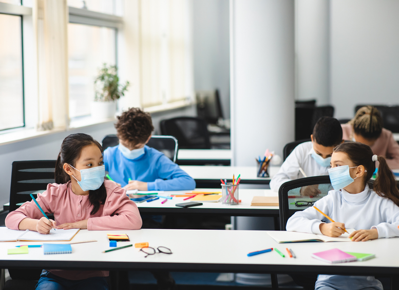 Niñas y niños en aula de clases sentados, con mascarillas, unos hablando y otros escribiendo