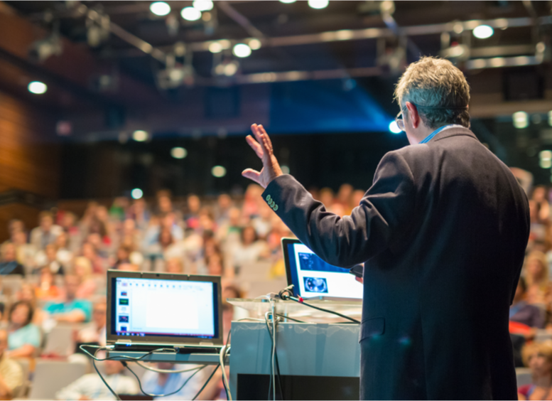 Conferencista de espaldas, hombre, en un auditorio lleno de personas, con su mano levantada, de pie y dos computadores de apoyo se dirige al público