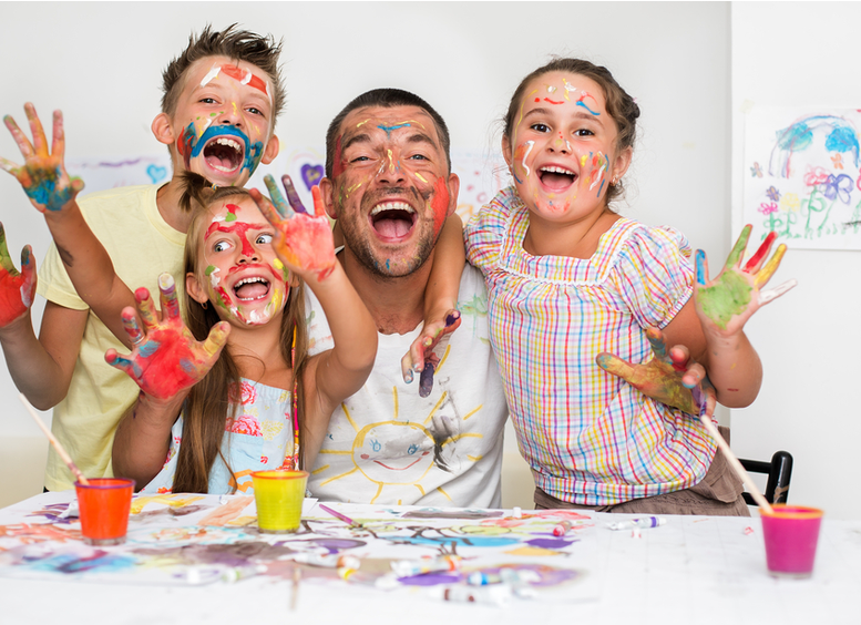 Padre con sus tres hijos, con la cara pintada, sonrientes mostrando sus manos pintadas de colores brillantes