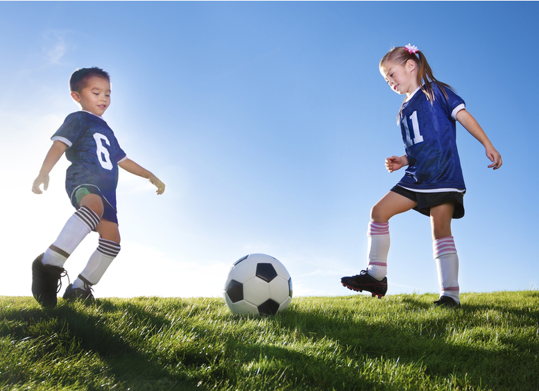 Niña y niño, con camiseta y pantaloneta azul, medias blancas, guayos, en un prado donde se encuentra el balòn blanco con rombos negros. Los dos niños mueven las piernas para pegarle al balón