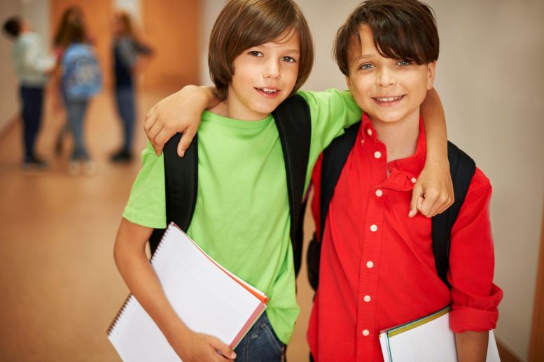 Dos niños en el colegio con cuadernos, pasan el brazo sobre el hombro del otro y sonríen a la cámara en un pasillo