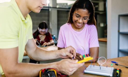 Dos estudiantes, una niña trigueña de blusa rosada y un joven de camiseta verde, aprendiendo circuitos electrónicos en clase de tecnología