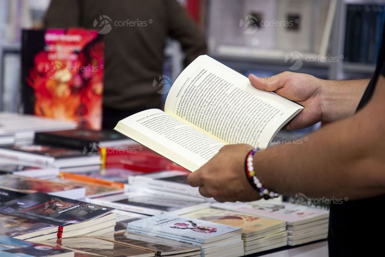 Foto en primer plano de unas manos sosteniendo un libro abierto, y muchos libros sobre una mesa